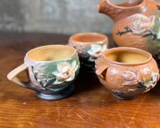 This lot features a collection of seven (7) Roseville Pottery items in the "Magnolia" pattern, which was introduced in 1943. The set includes a large cider pitcher and several matching mugs, featuring pink and white blossoms on a textured russet and green ground. All items are in excellent condition with one piece showing a very small chip as shown in photos. The pitcher measures, 8.5” H.
