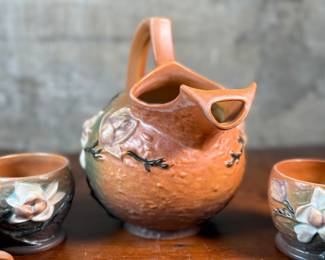 This lot features a collection of seven (7) Roseville Pottery items in the "Magnolia" pattern, which was introduced in 1943. The set includes a large cider pitcher and several matching mugs, featuring pink and white blossoms on a textured russet and green ground. All items are in excellent condition with one piece showing a very small chip as shown in photos. The pitcher measures, 8.5” H.
