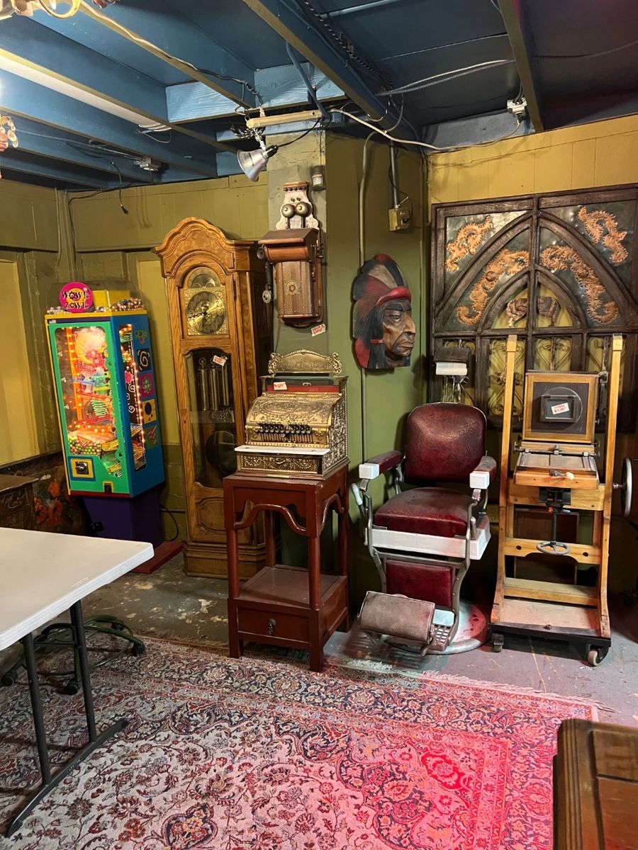 Vintage and antique interior vignette featuring a diverse assortment of collectible furnishings and display pieces. Includes an early coin-operated or trade stimulator–style machine with colorful painted front, an ornate wooden clock case, a small roll-top or writing cabinet, and an antique barber chair with adjustable metal arms and footrest upholstered in worn red leather. Also present is a carved architectural panel with Gothic arch detailing, a wall-mounted mask, and assorted wooden display stands and equipment. Pieces show visible age, wear, and patina throughout, consistent with long-term use and display. Ideal for collectors, decorators, set designers, or restoration projects. Sold as-is.