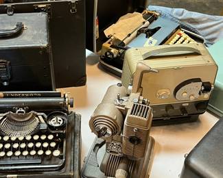 Vintage office and media equipment grouping featuring early typewriters, a reel-to-reel style projector or film device, and assorted mechanical and electrical components. Showcases classic mid-century industrial design with visible wear and patina. Ideal for collectors, display, or restoration projects. Functionality untested. All items sold as-is.