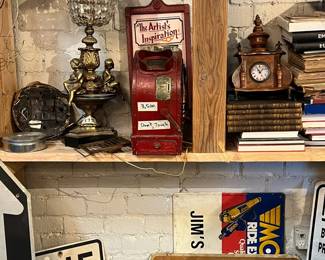 Vintage shelf display featuring an ornate table lamp with glass prisms, an early advertising or novelty coin-operated machine, antique mantel clock, stacked books, and assorted signage including street and parking signs. A rich mix of lighting, ephemera, and decorative objects with strong patina and old-world character. Ideal for collectors, decorators, or retail display. All items sold as-is.