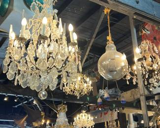 Impressive selection of antique and vintage crystal chandeliers and glass lighting fixtures hanging overhead. Features a variety of styles with cascading prisms, globe shades, and ornate metalwork, creating a luminous, old-world ambiance. Ideal for collectors, designers, or architectural lighting projects. All items sold as-is.