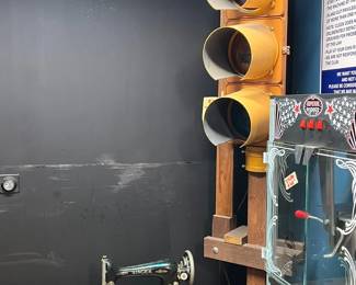 Industrial-style vignette featuring a vintage traffic signal mounted on a wooden stand, paired with an antique treadle-style sewing machine set into a wooden table. Also includes a glass-front display cabinet with mechanical or game-style elements. A bold mix of industrial and utilitarian design with strong visual impact. Ideal for collectors, decorators, or retail display. All items sold as-is.