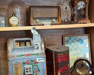 Vintage shelf display featuring a classic tabletop radio, novelty slot machine bank, decorative tins, early clocks, and assorted small collectibles. A charming mix of mechanical, advertising, and mid-century pieces with visible age and patina. Ideal for collectors, game rooms, or nostalgic décor. All items sold as-is.