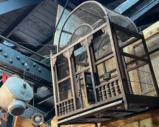 Large antique birdcage with arched wire top and wooden frame, featuring turned wood interior elements and original wire panels. Displayed overhead, showing extensive age, wear, and patina consistent with long-term use. A striking decorative or architectural accent ideal for collectors, garden display, or theatrical and retail staging. All items sold as-is.