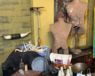 Eclectic antique and vintage décor grouping featuring a collection of decorative objects and curiosities. Includes two early dressmaker’s mannequins with original fabric forms on metal stands, showing age, wear, and surface distress consistent with long-term use. Also present are assorted decorative items such as antlers, horns, vintage bottles, barware, wood boxes, sculptural metal pieces, lighting elements, and assorted tabletop accessories. A carved wall plaque with a figural motif is visible in the background. Materials include wood, metal, fabric, glass, and mixed media. Ideal for decorators, collectors, theatrical props, or retail display. Condition varies throughout; all items show age-related wear. Sold as-is.