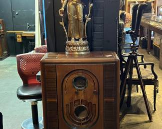 Vintage wooden floor radio cabinet featuring a richly grained case with rounded front, decorative inlay, and original circular dial and speaker openings. The cabinet stands on short legs and retains its classic Art Deco–era styling. Radio components appear present but functionality is untested. Topped with a decorative gilt metal standing deity sculpture (Asian-inspired, likely Thai or Hindu influence), adding visual interest for display or staging purposes. Cabinet shows age-appropriate wear, surface scratches, and patina consistent with vintage use. Ideal as a restoration project, decorative furniture piece, or statement prop. Sold as-is.