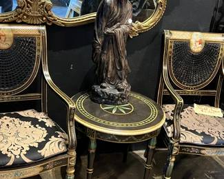 Refined antique vignette featuring a carved figural sculpture displayed atop a small round inlaid accent table, flanked by a pair of decorated side chairs with painted and gilt detailing and patterned upholstery. Set against an ornate gilt-framed mirror, the grouping reflects classic European-style elegance with age-appropriate wear and patina. Ideal for collectors or decorative display. All items sold as-is.
