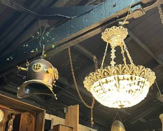 Dramatic antique lighting display featuring an ornate crystal chandelier with gilt framework alongside a hanging brass deep-sea diver’s helmet and vintage industrial elements. Suspended above shelves of early radios and collectibles, the scene offers bold contrast and strong visual impact. Ideal for collectors, designers, or statement interiors. All items sold as-is.