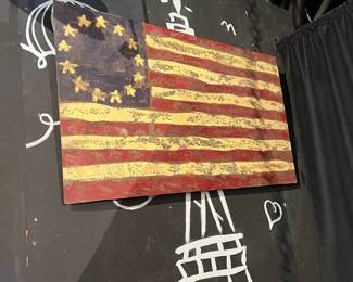 Vintage-style metal American flag wall sign with distressed, weathered finish. Features a rustic red, white, and blue design with a worn patina for an industrial or farmhouse look. Suitable for wall display in home, garage, or commercial décor.
