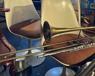 Vintage brass trombone displayed alongside mid-century modern molded chairs. Features classic brass construction with visible patina and wear consistent with age. A striking crossover piece suitable for musicians, collectors, or decorative display. Functionality untested. All items sold as-is.