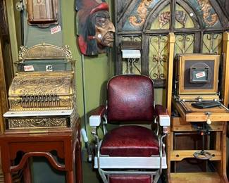 Striking antique display featuring an ornate early cash register, a vintage barber chair with red leather upholstery and adjustable metal arms, and an early photographic camera on stand. Accented by a carved architectural panel with Gothic-style detailing and wall-mounted decorative elements. Rich patina and strong period character throughout. Ideal for collectors, decorators, or theatrical display. All items sold as-is.