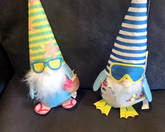 These are a set of two summer-themed decorative gnomes. 
The gnomes are plush and weighted for stability. 
One gnome wears a Hawaiian-print hat, sunglasses, and flip-flops, holding a coconut drink. 
