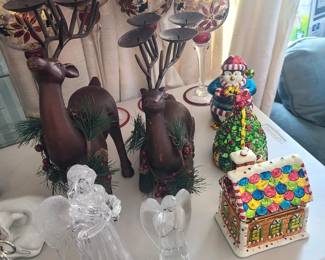 This image displays a collection of assorted Christmas decorations, including reindeer figures, candle holders, and figurines.
The collection features two large brown reindeer figures adorned with artificial pine sprigs and berries. 
There are four wine glass candle holders with decorative, gold-leaf-style bowls and dark metal candle dishes. 
Other items include two clear glass or acrylic angel figurines, a small gingerbread house decoration, and a snowman figure. 

