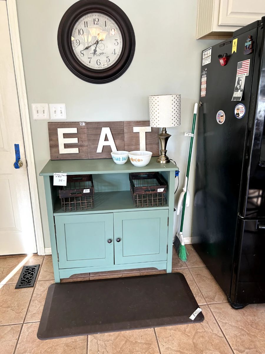 green cabinet, cloct two Pyrex bowls, lamp and three wall pictures spelling eat, cushion rug.