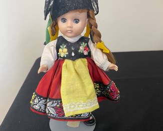 a vintage Vogue Ginny Doll from the "Far Away Lands" collection, representing Switzerland.
The 8-inch vinyl doll wears a traditional Swiss costume with a red skirt, white blouse, black vest with floral embroidery, and a yellow apron.
