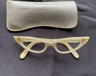 a pair of vintage cat-eyeglasses, a popular style from the 1950s and 1960s, The frames are a light color with small decorative floral or leaf-like embellishments near the temples.
