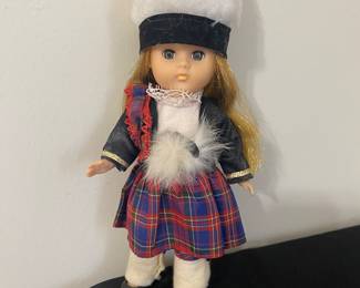 vintage Vogue Ginny doll. Wears a traditional Scottish outfit including a red plaid kilt, a black jacket with gold trim, a white blouse with lace, white leggings, and a white plush sporran (pouch).