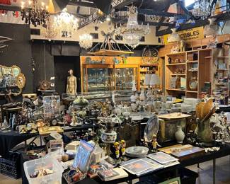 Wide interior view of estate sale showcasing a large volume of antiques, collectibles, and decorative objects. Tables filled with glassware, art, smalls, and curios, with chandeliers and lighting overhead. Displays highlight the depth, variety, and quantity available throughout the sale.