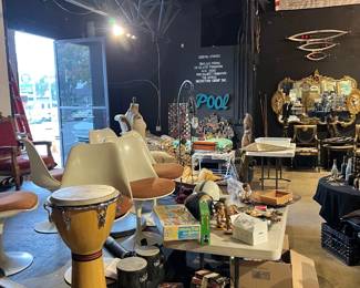 Another wide view of the estate sale floor featuring furniture, lighting, musical instruments, and assorted collectibles. Includes seating, décor, boxed items, and display tables arranged throughout the space. Emphasizes the extensive selection and diverse categories available at the sale.