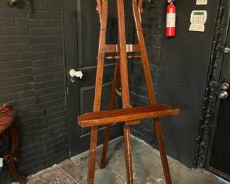 Ornate vintage floor-standing easel with carved wood construction and decorative scrollwork. Features an adjustable support and rich, dark finish with gilt-style accents. Ideal for displaying artwork, mirrors, or signage in a gallery, studio, or decorative interior.
