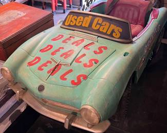 Vintage amusement ride car with hand-painted “Used Cars / Deals Deals Deals” signage. Features a whimsical, weathered green body with red interior seating and charming folk-art character. Likely from a carnival or kiddie ride attraction. Eye-catching decorative piece ideal for commercial display, retail décor, or themed interiors.