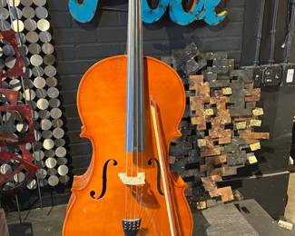Full-size cello with warm amber-toned varnish and classic f-hole design, displayed on a floor stand. Includes bow. Traditional acoustic string instrument suitable for players, students, or decorative studio display. Shows light wear consistent with use.