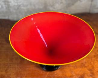 This lot features a stunning postmodern art glass bowl by IBEX Glass Studio circa 1992. This bowl is in excellent condition, signed underneath, and measures, 8” H x 13” W x 13” D. Formed in 1985 by Dimitri Michaelides, Sam Stang and David Levi, IBEX Glass Studio was heavily influenced by American studio glass pioneers, mid-century Venetian glass, and postmodern European design, including Ettore Sottsass and his Memphis contemporaries. Sitting on a round black foot, this bowl has a tapered cylindrical red bowl, and a delicate yellow rim.

