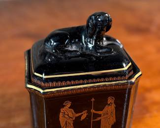 This lot features an antique English tea caddy, attributed to Henry Clay circa 1850s. It features classical Etruscan-style figures and borders with the top decorated with a reclining dog figure. This caddy measures, 5.5” H x 4.5” W x 3.5” D.

