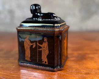 This lot features an antique English tea caddy, attributed to Henry Clay circa 1850s. It features classical Etruscan-style figures and borders with the top decorated with a reclining dog figure. This caddy measures, 5.5” H x 4.5” W x 3.5” D.

