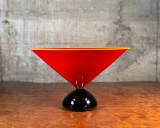 This lot features a stunning postmodern art glass bowl by IBEX Glass Studio circa 1992. This bowl is in excellent condition, signed underneath, and measures, 8” H x 13” W x 13” D. Formed in 1985 by Dimitri Michaelides, Sam Stang and David Levi, IBEX Glass Studio was heavily influenced by American studio glass pioneers, mid-century Venetian glass, and postmodern European design, including Ettore Sottsass and his Memphis contemporaries. Sitting on a round black foot, this bowl has a tapered cylindrical red bowl, and a delicate yellow rim.

