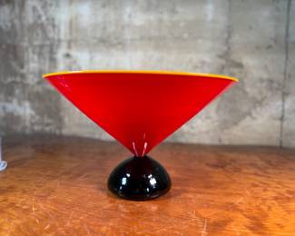 This lot features a stunning postmodern art glass bowl by IBEX Glass Studio circa 1992. This bowl is in excellent condition, signed underneath, and measures, 8” H x 13” W x 13” D. Formed in 1985 by Dimitri Michaelides, Sam Stang and David Levi, IBEX Glass Studio was heavily influenced by American studio glass pioneers, mid-century Venetian glass, and postmodern European design, including Ettore Sottsass and his Memphis contemporaries. Sitting on a round black foot, this bowl has a tapered cylindrical red bowl, and a delicate yellow rim.

