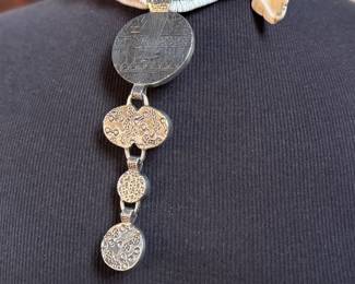 This lot features a special custom necklace by listed designer, Melanie DeLuca. Engraved sterling silver with shells & stones throughout. This piece measures, 17” and has a total approx. weight of 193.49 grams. Melanie DeLuca’s bold jewelry designs of mixed media, plastics and ethnographic objects embody a celebration of her 45 years as a metalsmith, jewelry designer and master teacher. Her interest in jewelry design began early and solidified at 17. By 20, she was apprenticing for master silversmith Brooks Darrow (himself an apprentice of renowned English architect and master metalsmith Louis Osman). Early work in painting and sculpture informed Melanie’s growing connection to the limitless expression of metal through jewelry design and fabrication.

