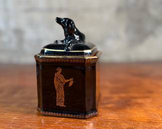 This lot features an antique English tea caddy, attributed to Henry Clay circa 1850s. It features classical Etruscan-style figures and borders with the top decorated with a reclining dog figure. This caddy measures, 5.5” H x 4.5” W x 3.5” D.


