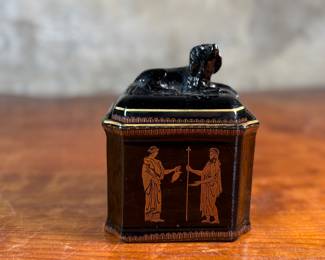 This lot features an antique English tea caddy, attributed to Henry Clay circa 1850s. It features classical Etruscan-style figures and borders with the top decorated with a reclining dog figure. This caddy measures, 5.5” H x 4.5” W x 3.5” D.


