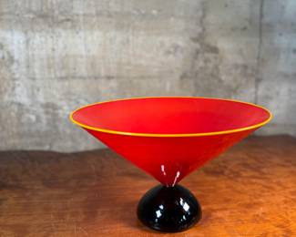 This lot features a stunning postmodern art glass bowl by IBEX Glass Studio circa 1992. This bowl is in excellent condition, signed underneath, and measures, 8” H x 13” W x 13” D. Formed in 1985 by Dimitri Michaelides, Sam Stang and David Levi, IBEX Glass Studio was heavily influenced by American studio glass pioneers, mid-century Venetian glass, and postmodern European design, including Ettore Sottsass and his Memphis contemporaries. Sitting on a round black foot, this bowl has a tapered cylindrical red bowl, and a delicate yellow rim.

