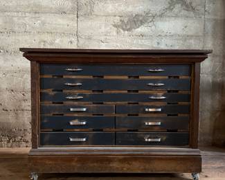 This lot features a vintage solid wood apothecary style or print maker’s cabinet. Comes on casters with multiple sized pull out drawers. The top "lift up" board is currently not attached to the backside trim, but can be easily fixed or left as is. The top cabinet area has green felt. This cabinet measures, 33.25” H x 46” W x 23” D.

