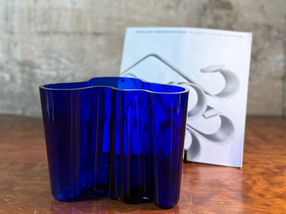 This lot features a Iittala Alvar Aalto Collection vase in ultramarine blue mouth-blown glass. It is also known as the Savoy Vase. The vase was designed by Finnish architect Alvar Aalto and his wife Aino for a design competition in 1936. It became an iconic piece of Finnish design after being displayed at the 1937 Paris World’s Fair. The unique, wavy shape was reportedly inspired by the hem of a Sami woman’s dress, with original sketches titled “The Eskimo Woman’s Leather Breeches”. Each vase is still manufactured by skilled glassblowers at the Iittala factory in Finland using original wooden molds. This vase is accompanied with Aalto’s MOMA book.


