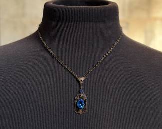 This lot features an antique Art Deco sterling silver filigree pendant necklace with a large blue central stone, likely blue chalcedony or glass, and marcasite accents. The intricate filigree design and geometric patterns are characteristic of the Art Deco period, circa the 1920s-1930s. This necklace measures, 18”.


