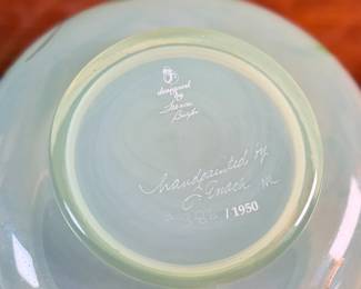 This lot features a Fenton "Dahlias" on Topaz Opalescent covered box, hand-painted by artist Frances Burton. Edition 386/1950 and measures, 5.5” H x 7” W x 7” D. The piece is crafted from Topaz Opalescent glass, which is a yellow mist color created by striking the opalescent glass before forming. The dahlia design features blue flowers with 22k gold highlights. The glass is also known as Vaseline glass and glows under a blacklight due to the use of depleted uranium in the glassmaking process.

