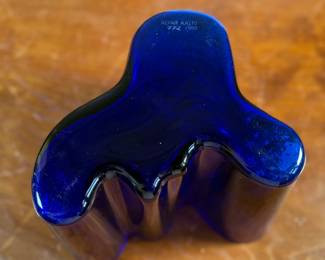 This lot features a Iittala Alvar Aalto Collection vase in ultramarine blue mouth-blown glass. It is also known as the Savoy Vase. The vase was designed by Finnish architect Alvar Aalto and his wife Aino for a design competition in 1936. It became an iconic piece of Finnish design after being displayed at the 1937 Paris World’s Fair. The unique, wavy shape was reportedly inspired by the hem of a Sami woman’s dress, with original sketches titled “The Eskimo Woman’s Leather Breeches”. Each vase is still manufactured by skilled glassblowers at the Iittala factory in Finland using original wooden molds. This vase is accompanied with Aalto’s MOMA book.

