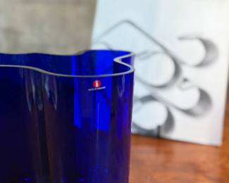 This lot features a Iittala Alvar Aalto Collection vase in ultramarine blue mouth-blown glass. It is also known as the Savoy Vase. The vase was designed by Finnish architect Alvar Aalto and his wife Aino for a design competition in 1936. It became an iconic piece of Finnish design after being displayed at the 1937 Paris World’s Fair. The unique, wavy shape was reportedly inspired by the hem of a Sami woman’s dress, with original sketches titled “The Eskimo Woman’s Leather Breeches”. Each vase is still manufactured by skilled glassblowers at the Iittala factory in Finland using original wooden molds. This vase is accompanied with Aalto’s MOMA book.

