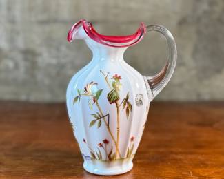 This lot features a Fenton Art Glass "Asian Garden" pitcher from the 2000 Connoisseur Collection. Limited edition 894/1850 and measures, 7.5” H x 5” W x 6” D. It features a hand-painted "Asian Garden" design on milk glass with a peach crest and a clear applied handle. The design was created by artist Robin Spindler and includes 22k gold highlights. 

