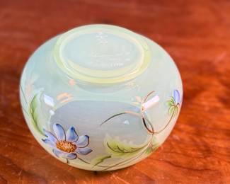 This lot features a Fenton "Dahlias" on Topaz Opalescent covered box, hand-painted by artist Frances Burton. Edition 386/1950 and measures, 5.5” H x 7” W x 7” D. The piece is crafted from Topaz Opalescent glass, which is a yellow mist color created by striking the opalescent glass before forming. The dahlia design features blue flowers with 22k gold highlights. The glass is also known as Vaseline glass and glows under a blacklight due to the use of depleted uranium in the glassmaking process.

