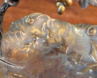 This lot features an American Aesthetic Movement trompe l’oeil basket, naturalistically modeled to simulate a cloth dish with a handle made of a bent twig and corded twine. The basket features gilt and silvered leaves and flower heads as decoration. The piece dates back to the 1940s–1950s and is made of solid brass. This piece measures, 7.5” H x 7.75” W x 7.75” D.

