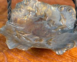 This lot features an American Aesthetic Movement trompe l’oeil basket, naturalistically modeled to simulate a cloth dish with a handle made of a bent twig and corded twine. The basket features gilt and silvered leaves and flower heads as decoration. The piece dates back to the 1940s–1950s and is made of solid brass. This piece measures, 7.5” H x 7.75” W x 7.75” D.

