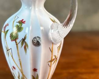 This lot features a Fenton Art Glass "Asian Garden" pitcher from the 2000 Connoisseur Collection. Limited edition 894/1850 and measures, 7.5” H x 5” W x 6” D. It features a hand-painted "Asian Garden" design on milk glass with a peach crest and a clear applied handle. The design was created by artist Robin Spindler and includes 22k gold highlights. 

