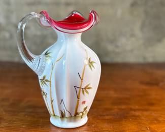 This lot features a Fenton Art Glass "Asian Garden" pitcher from the 2000 Connoisseur Collection. Limited edition 894/1850 and measures, 7.5” H x 5” W x 6” D. It features a hand-painted "Asian Garden" design on milk glass with a peach crest and a clear applied handle. The design was created by artist Robin Spindler and includes 22k gold highlights. 

