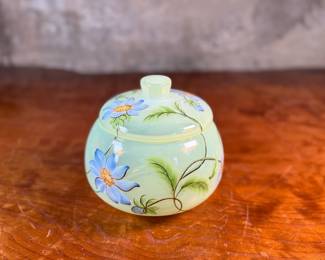 This lot features a Fenton "Dahlias" on Topaz Opalescent covered box, hand-painted by artist Frances Burton. Edition 386/1950 and measures, 5.5” H x 7” W x 7” D. The piece is crafted from Topaz Opalescent glass, which is a yellow mist color created by striking the opalescent glass before forming. The dahlia design features blue flowers with 22k gold highlights. The glass is also known as Vaseline glass and glows under a blacklight due to the use of depleted uranium in the glassmaking process.

