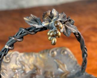 This lot features an American Aesthetic Movement trompe l’oeil basket, naturalistically modeled to simulate a cloth dish with a handle made of a bent twig and corded twine. The basket features gilt and silvered leaves and flower heads as decoration. The piece dates back to the 1940s–1950s and is made of solid brass. This piece measures, 7.5” H x 7.75” W x 7.75” D.


